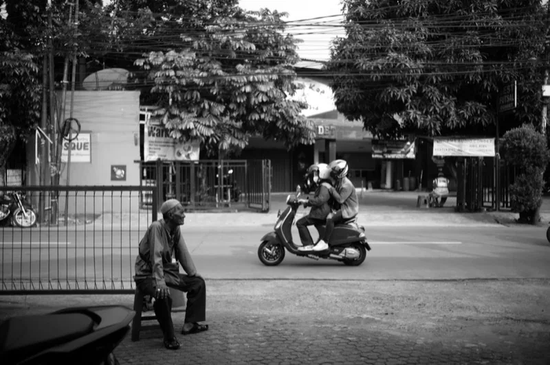 couple riding a motor scooter and an elderly man sitting at the gate of parking lot — illustrative editorial photography, not a verified location